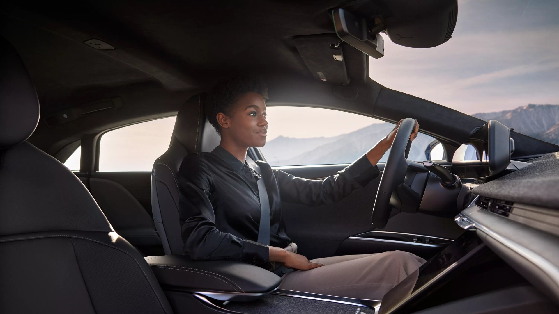 Une femme conduisant une Lucid Air affiche un air confiant et heureux. La photo est prise à partir de l'espace pour les jambes au siège du passager. Des montagnes sont visibles par les fenêtres de la voiture derrière la femme.