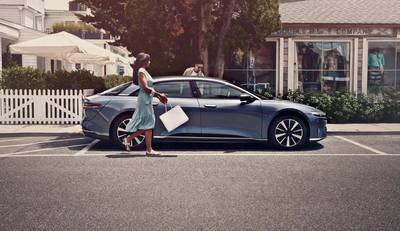 Een man en een vrouw stappen in een Fathom Blue Metallic Lucid Air Pure. De foto is gemaakt in een stranddorpje met winkels en parasols op de achtergrond.