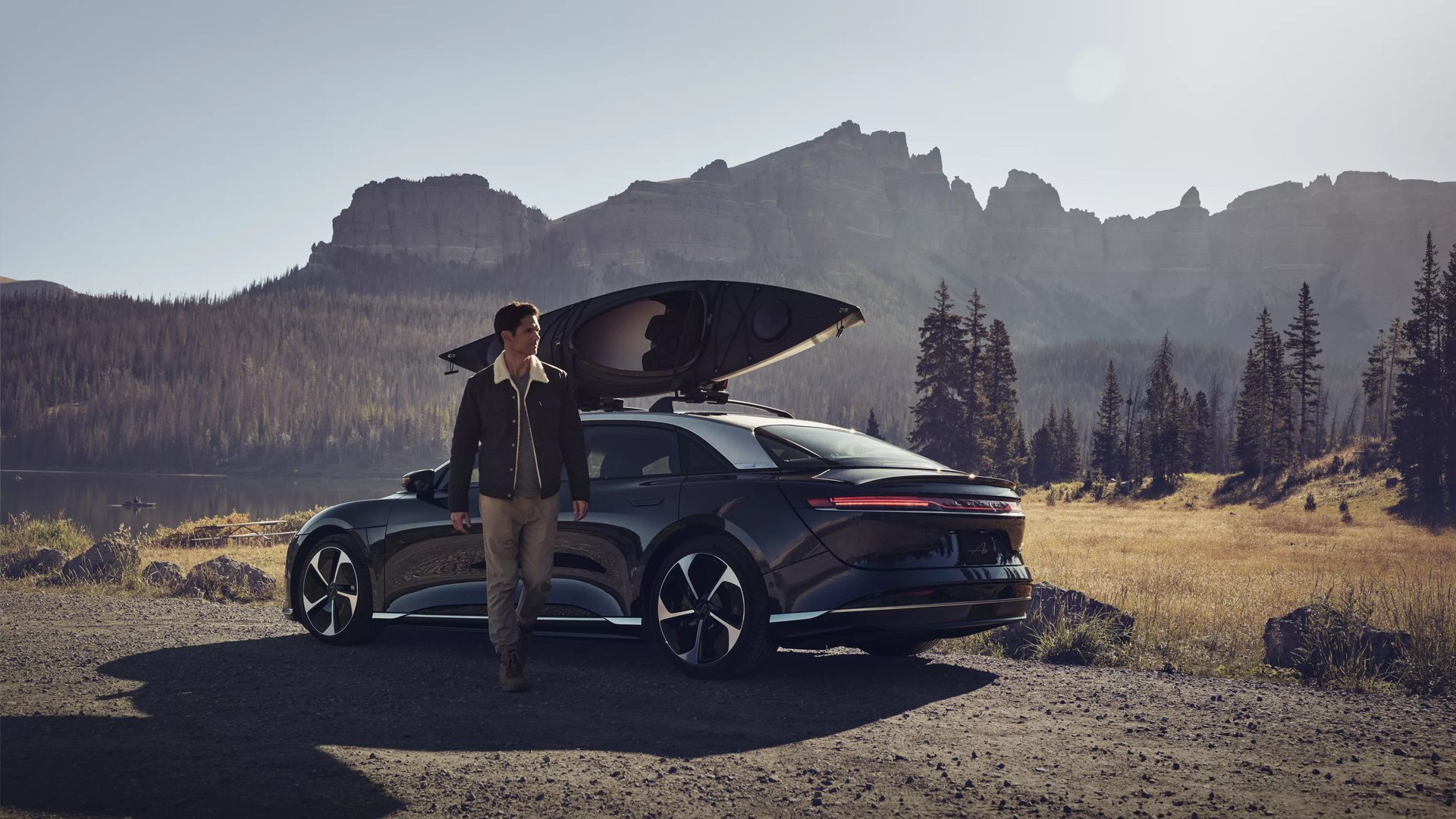 A man is walking towards the camera in front of a Infinite Black Metallic Lucid Air Pure. The Lucid Air is equipped with a roof carrier on which a cannoe is loaded. The background shows a lake, trees, and tall, rocky moutains.
