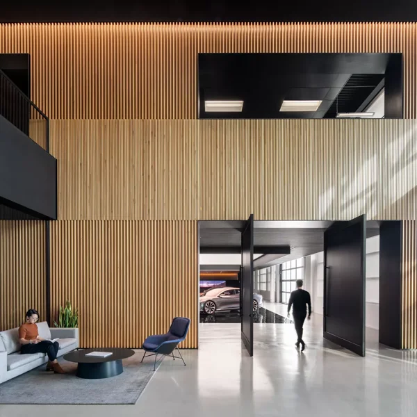 2 people, one sitting in a couch and ine walking away from the camera, are shown in a large space in a Lucid location. The room mixes wood and black colors, with concrete floors and lots of natural light.